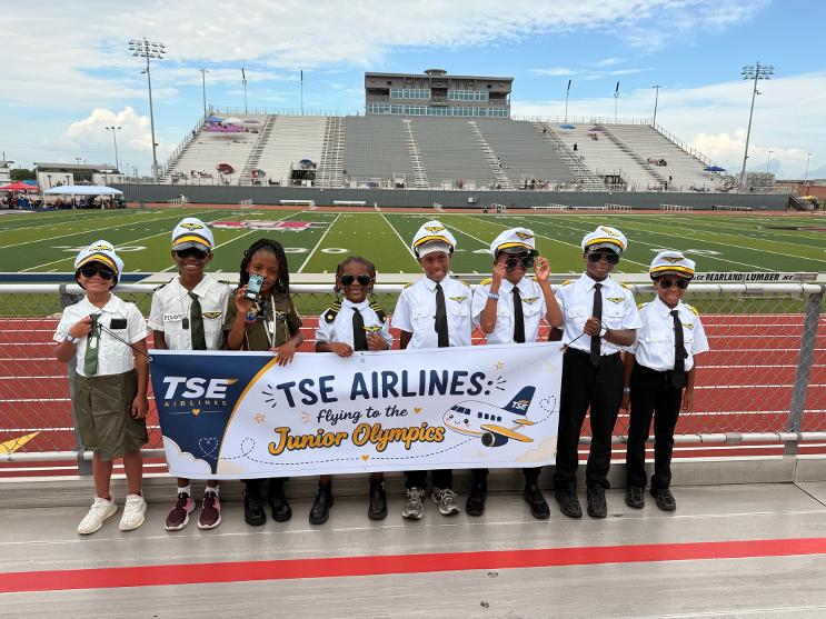 Young TSE athletes in pilot uniforms holding a TSE Airlines banner before the Junior Olympics
